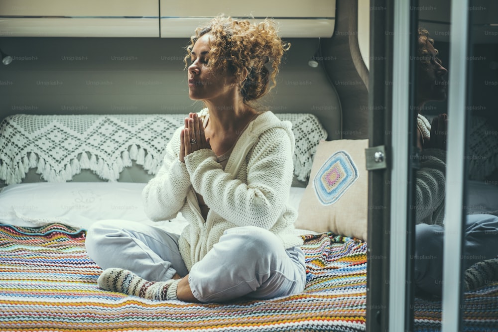 Young woman in meditation activity sitting on the bed in camper van bedroom. Serene and confident female people in yoga loto balance position. Healhty lifestyle lady praying and relaxing indoor