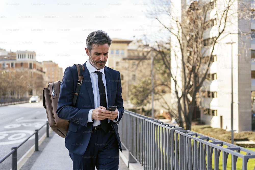 Empresário de meia-idade em terno formal navegando pelo celular enquanto caminha na cidade e se desloca para o trabalho