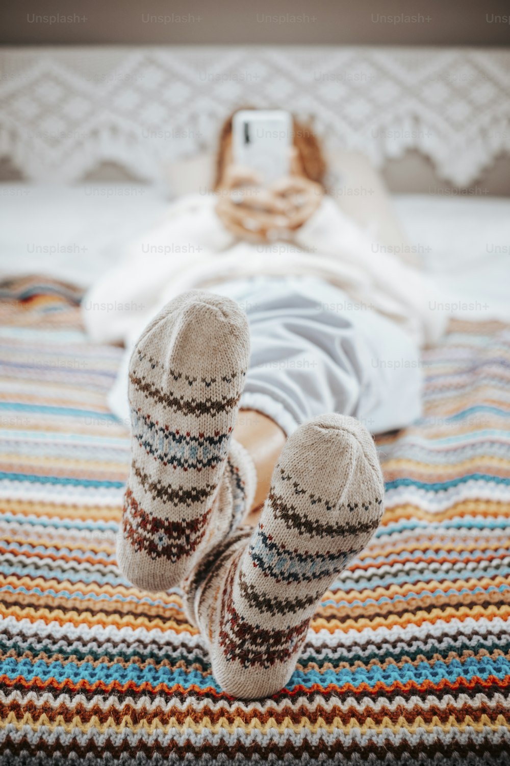 Close up of feet and nice socks on a same texture crochet woolen colorful blanket. Woman having relax leisure activity on the bedroom and use mobile phone in defocused background.