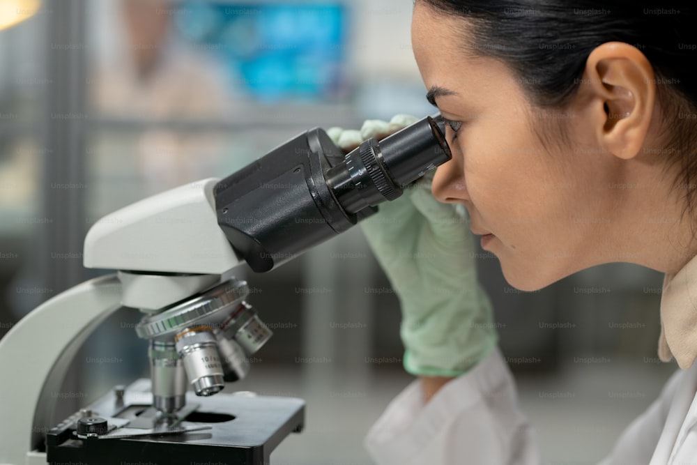Jeune femme scientifique en gants de protection regardant au microscope tout en étudiant une nouvelle substance chimique et ses caractéristiques