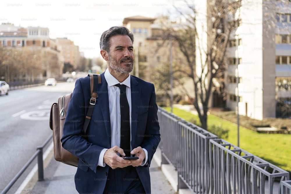 Empresario masculino de mediana edad en traje formal navegando por teléfono celular mientras camina por la ciudad y viaja al trabajo