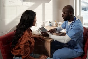 Junge selbstbewusste Ärztin, die auf den Tablet-Bildschirm zeigt, während sie einer Patientin während der medizinischen Konsultation ein neues Medikament präsentiert