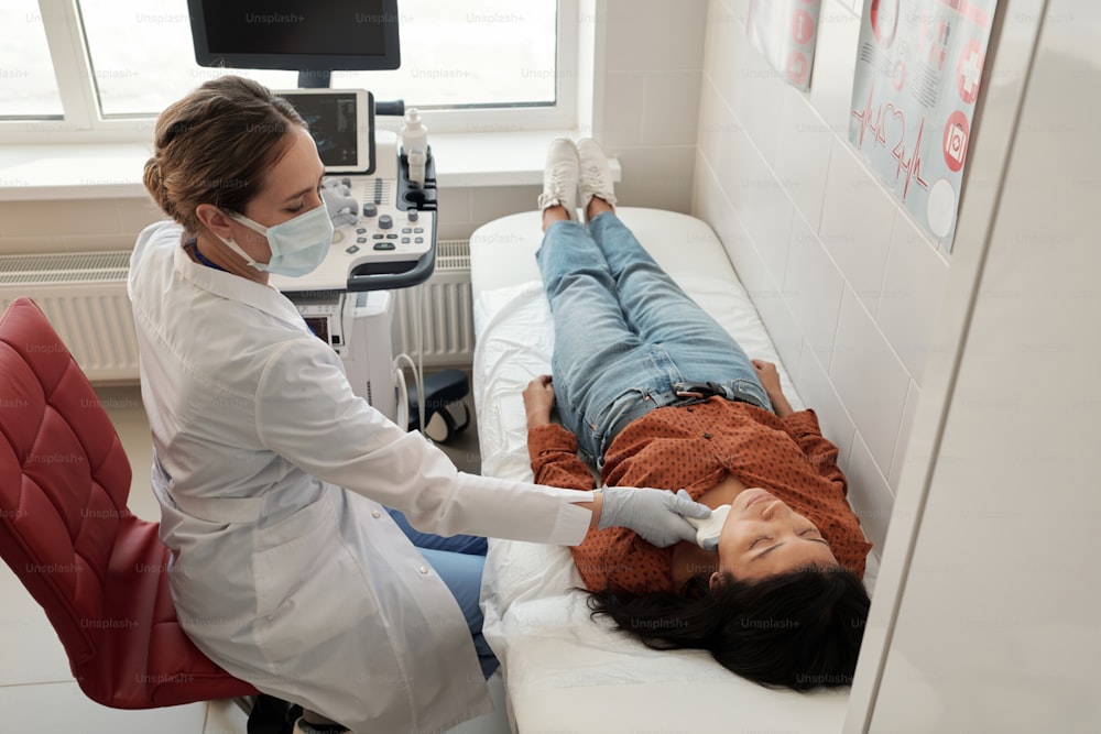 Zeitgenössischer junger Kliniker scannt Hals einer kranken hispanischen Patientin während der Ultraschalluntersuchung in Kliniken