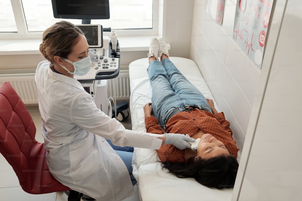 Contemporary young clinician scanning neck of sick Hispanic female patient during ultrasound examination in clinics