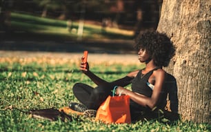 A side view of a beautiful curly-haired African female sitting on the lawn and leaning against a park tree while having a video call with her friend via a smartphone; a young black woman making selfie