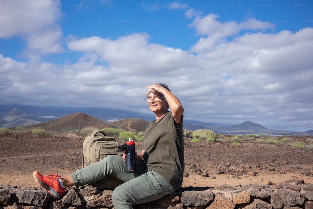 屋外ハイキングで石垣に座り、水筒を持って微笑む年配の女性。自由と晴れた日を楽しんでいる彼女のバックパックの近くに座っているアクティブな成熟した女性