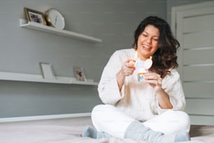 Young adult happy beautiful woman forty years plus size body positive in white cozy knitted sweater with festive cupcake with candle in hands on bed at the home party, birthday girl