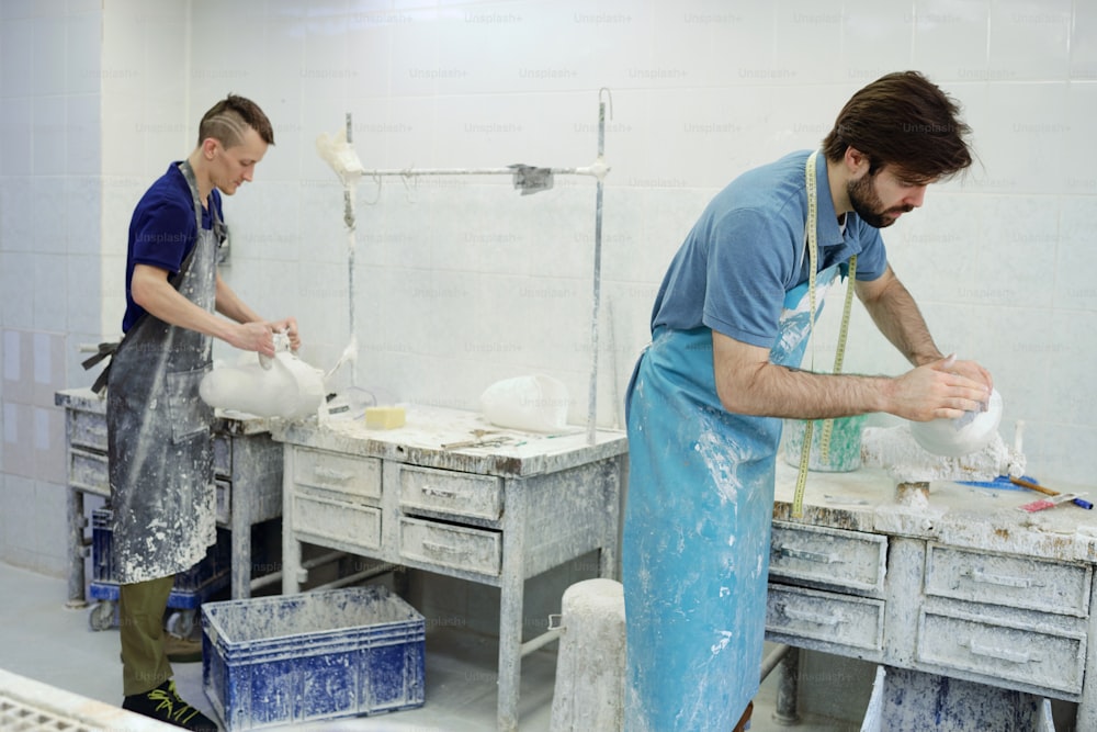 Two young male workers of prosthetic factory standing by tables while polishing plaster casts of residual limbs in workshop