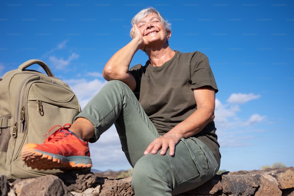Smiling elderly woman on outdoor hike sitting on a stone wall looking up to the sun. Active mature woman sitting near her backpack enjoying freedom and sunny day