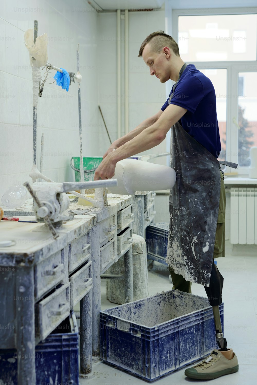 Young contemporary male worker in apron producing plaster workpiece of prostheic limb in workshop while standing by table