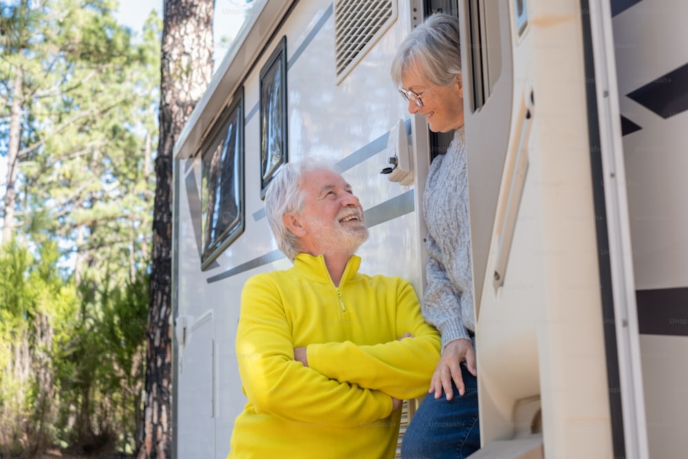 Carefree attractive senior couple standing out of their camper van motor home talking and smiling each other.  Elderly people enjoying freedom vacation travel in the forest