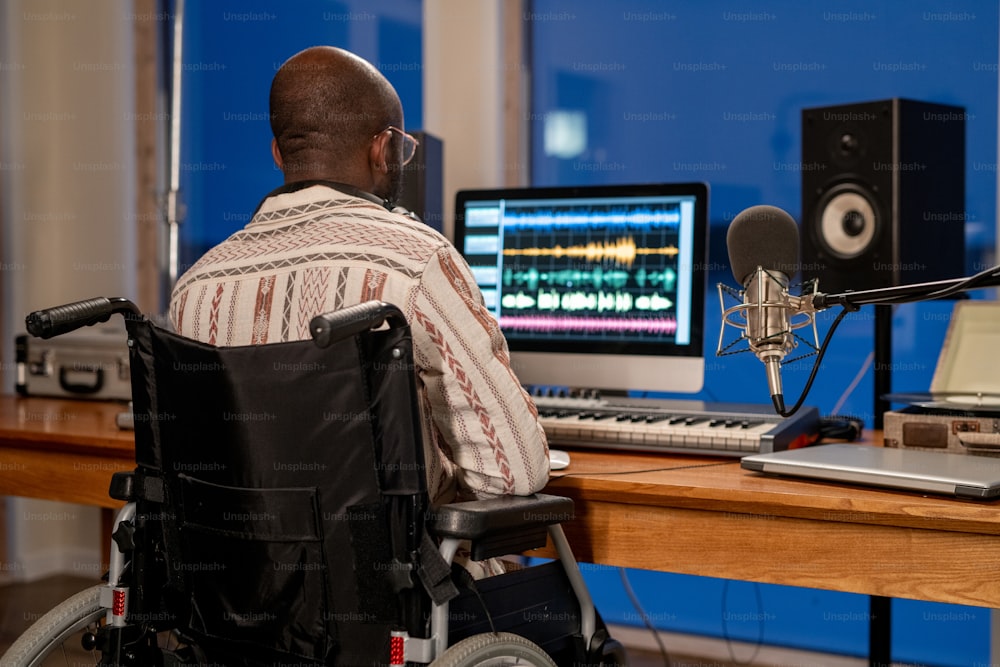 Rear view of man with disability using sound recording program while sitting by table in front of computer monitor in studio