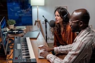 Junge Frau mit Kopfhörern singt im Mikrofon und schwarzer Mann nimmt ihre Lieder auf, während beide vor dem Computer sitzen