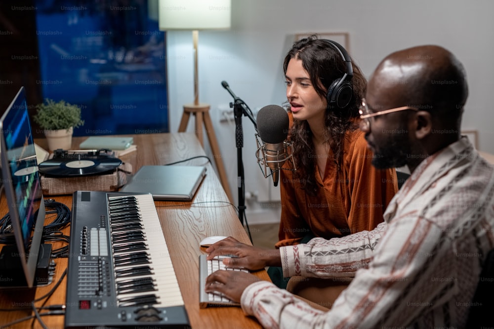 Young female in headphones singing in microphone and black man recording her songs while both sitting in front of computer