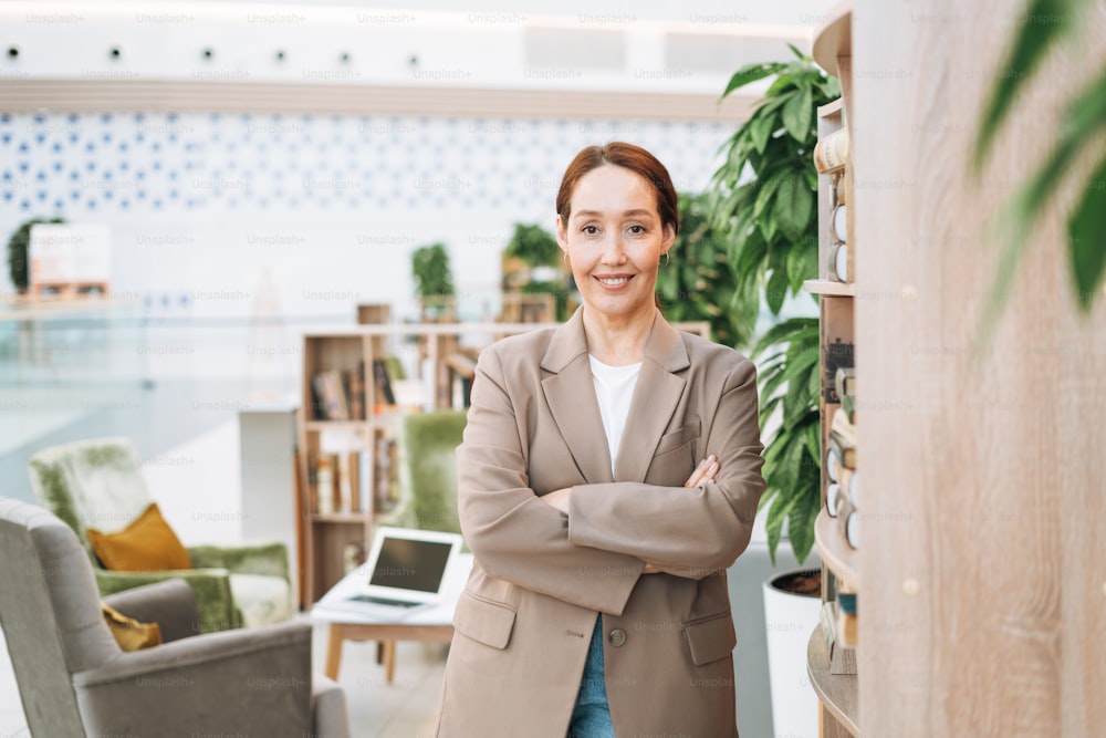 Adult smiling brunette business woman forty years with long hair in stylish beige suit and jeans at public place, green open space office, coworking. Friendly teacher or mentor with book in the library