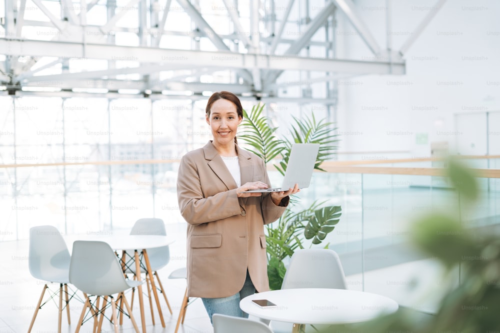 Adult smiling brunette business woman forty years with long hair in stylish beige suit and jeans working on laptop at the public place, open space office