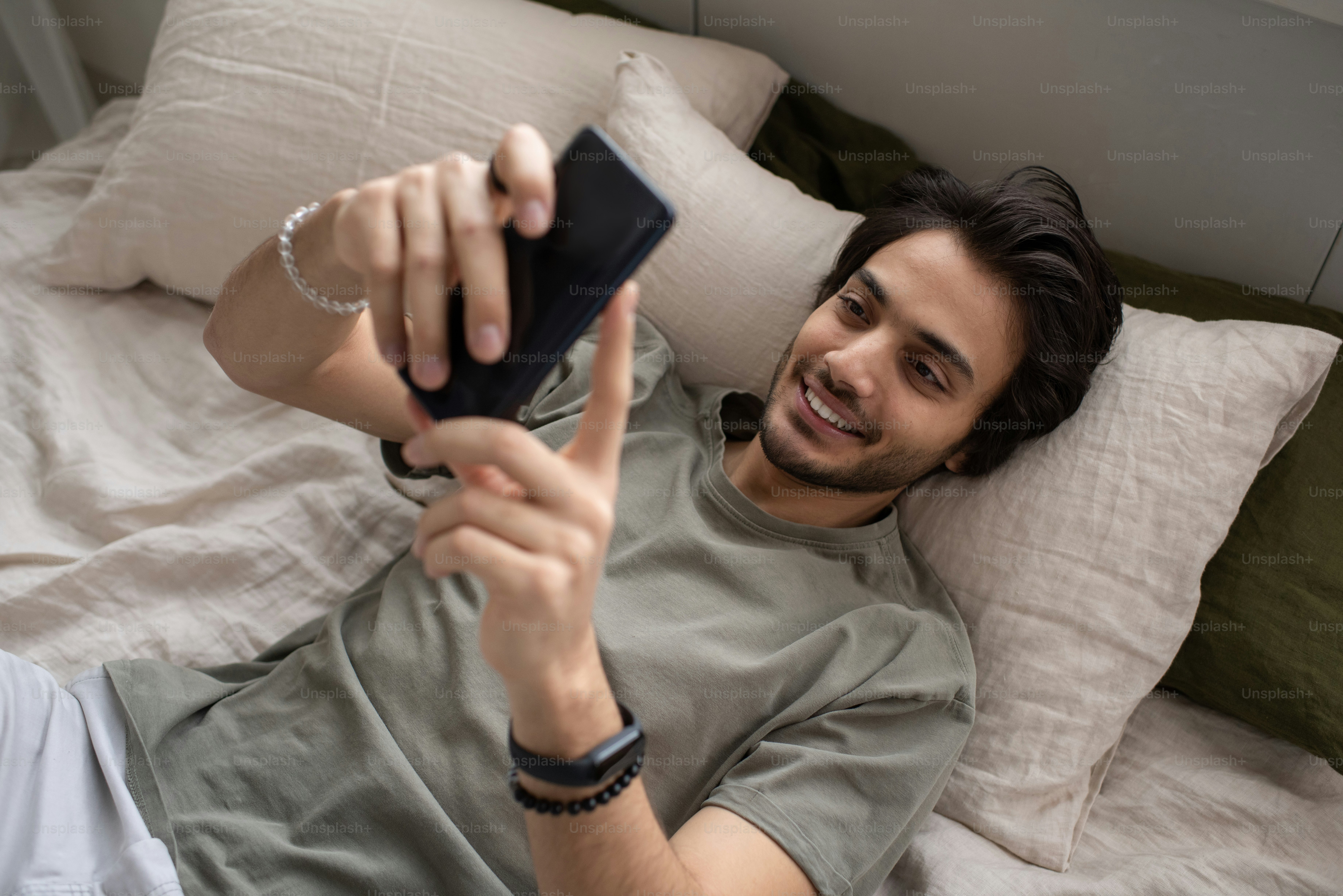 グレーのTシャツを着た若い笑顔の男性マネージャーがスマートフォンでテキストメッセージを送ったり、ベッドに横たわったままビデオ通話でコミュニケーションを取ったりする