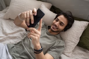 Young smiling male manager in grey t-shirt texting in smartphone or communicating through video call while lying on bed