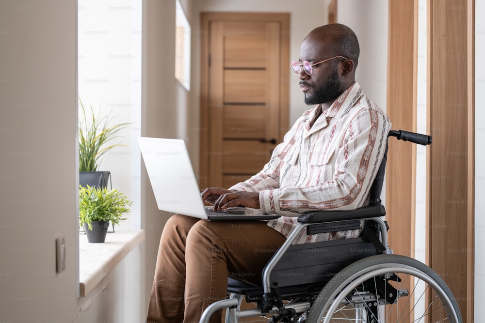 Giovane uomo nero serio con disabilità che si concentra sulla rete mentre si siede su una sedia a rotelle nel corridoio dell'appartamento