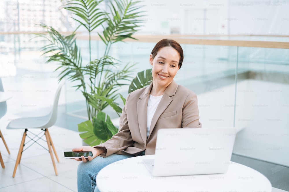 Femme d’affaires brune adulte souriante de quarante ans aux cheveux longs dans un élégant costume beige et un jean travaillant sur un ordinateur portable dans un lieu public, un bureau en open space