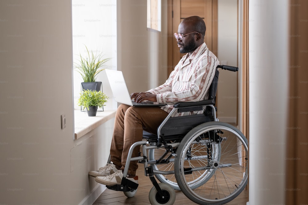 Giovane uomo nero serio in abbigliamento casual che digita sul computer portatile mentre è seduto in sedia a rotelle davanti alla finestra con piante verdi nel corridoio