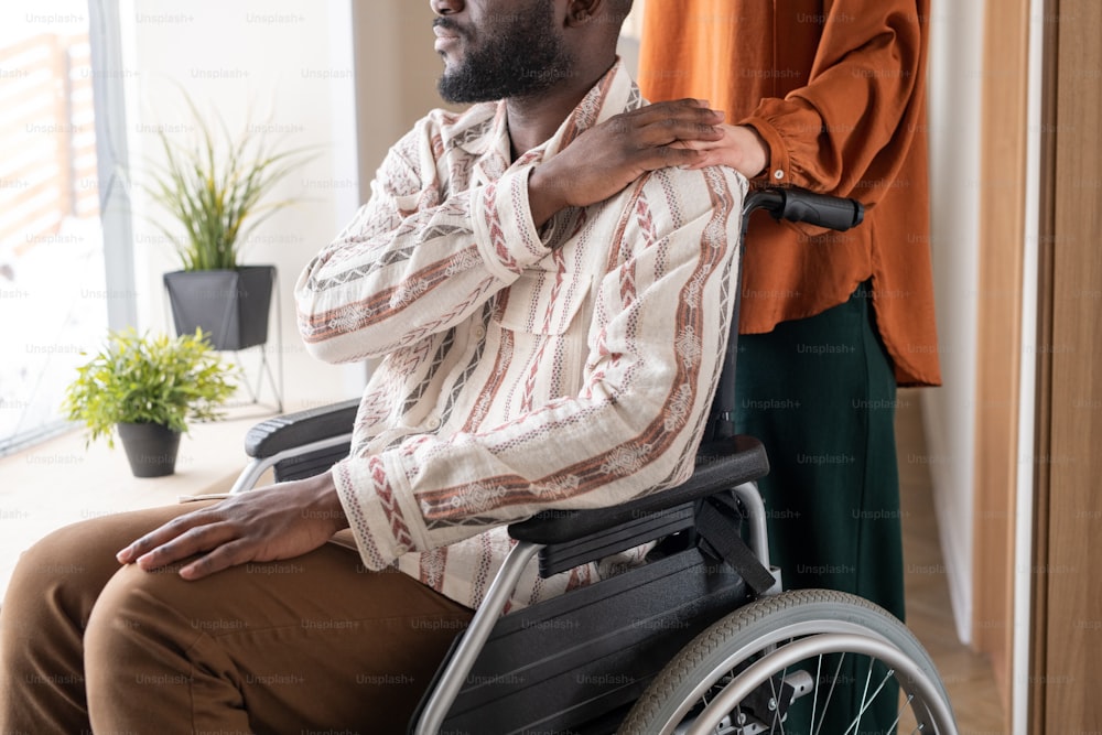 Side view of young African American man with disability keeping his hand on that of Caucasian female caregiver standing behind