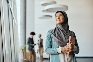 Mujer de negocios pensativa del Medio Oriente mirando a través de la ventana mientras está de pie en el pasillo de un edificio de oficinas.