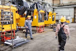 Blurred motion of two workers of industrial plant walking along spacious workshop with rows of huge machines and other equipment