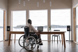 Hombre afroamericano con discapacidad trabajando frente a una computadora portátil mientras está sentado junto a la mesa frente a la ventana en la sala de estar