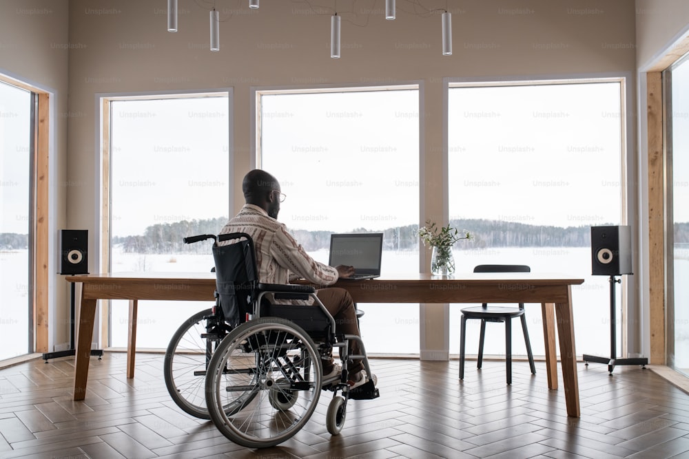 장애가 있는 아프리카계 미국인 남성은 거실의 랩지 창 앞 테이블에 앉아 노트북 앞에서 일하고 있다