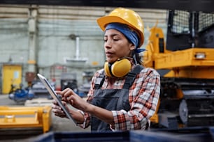 Young female technician in workwear scrolling through electronic document containing technical data in industrial workshop