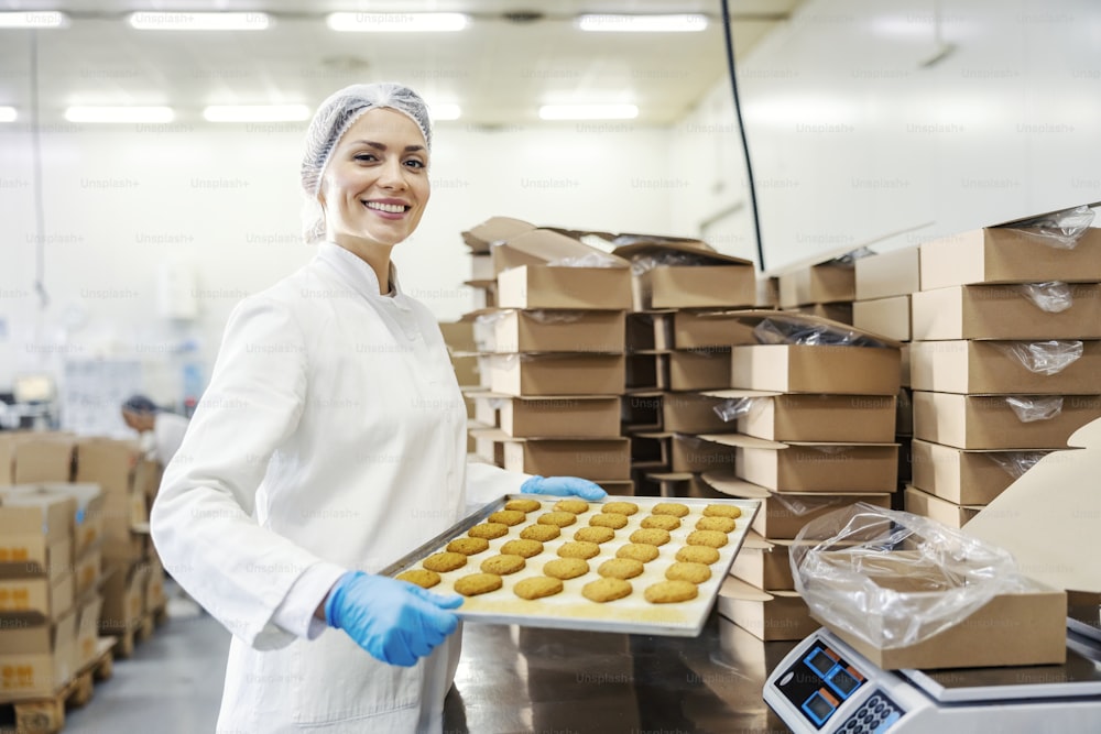 Glückliche Arbeiterin einer Lebensmittelfabrik, die ein Tablett mit Backkeksen hält und in die Kamera lächelt.