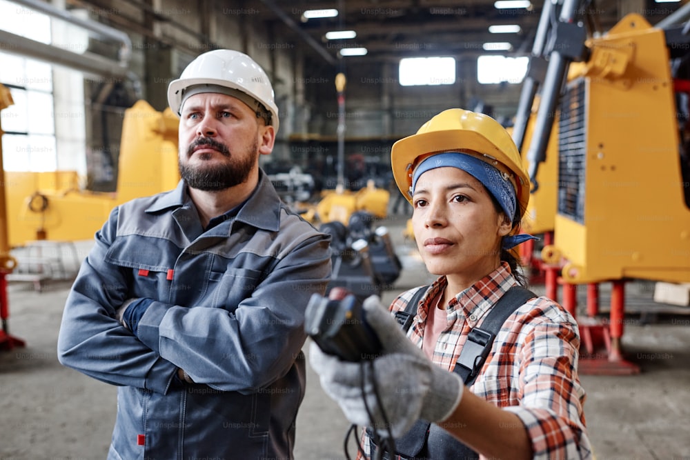 Dos trabajadores interraciales serios de una gran fábrica que miran una enorme máquina industrial mientras una técnica regula su altura