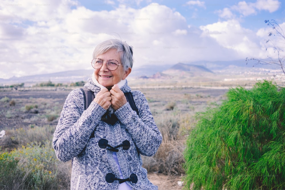 Attractive mature senior woman sitting alone outdoors with backpack on shoulders covering her neck with sweater. Smiling elderly woman enjoying freedom and travel, mountain and blue sky in background