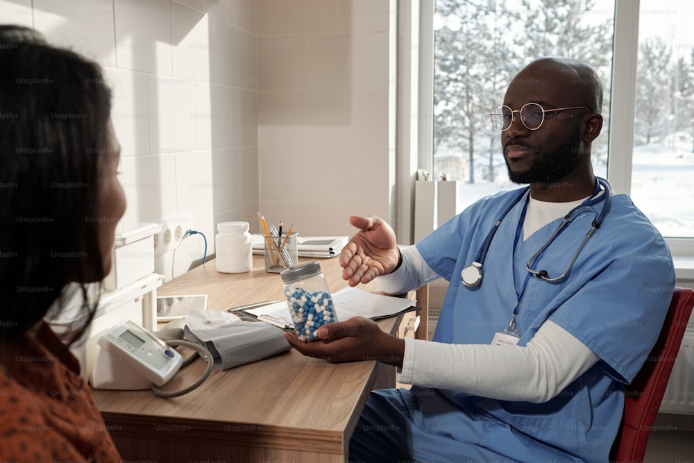 Junge afroamerikanische Klinikerin in blauer Uniform, die ein Glas mit Pillen hält und es einer Patientin im Krankenhaus empfiehlt