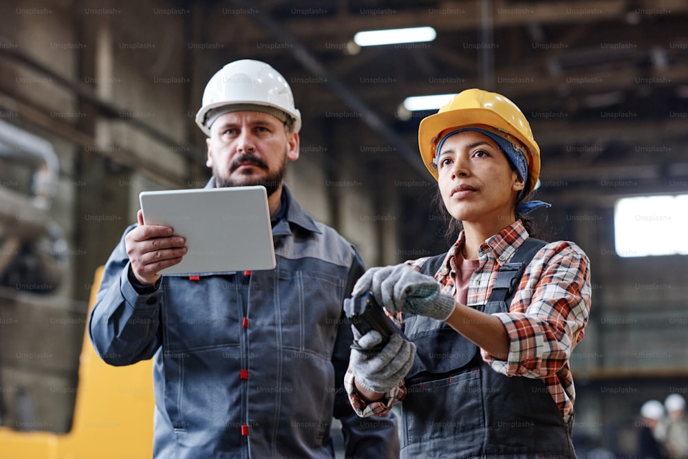 Dos ingenieros contemporáneos con tableta y control remoto mirando una enorme máquina industrial mientras una mujer regula su trabajo