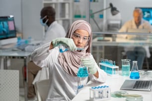 Junge ernsthafte muslimische Forscherin in Hijab, Handschuhen und Schutzbrillen gießt flüssige Substanz in Flasche mit blauer Flüssigkeit