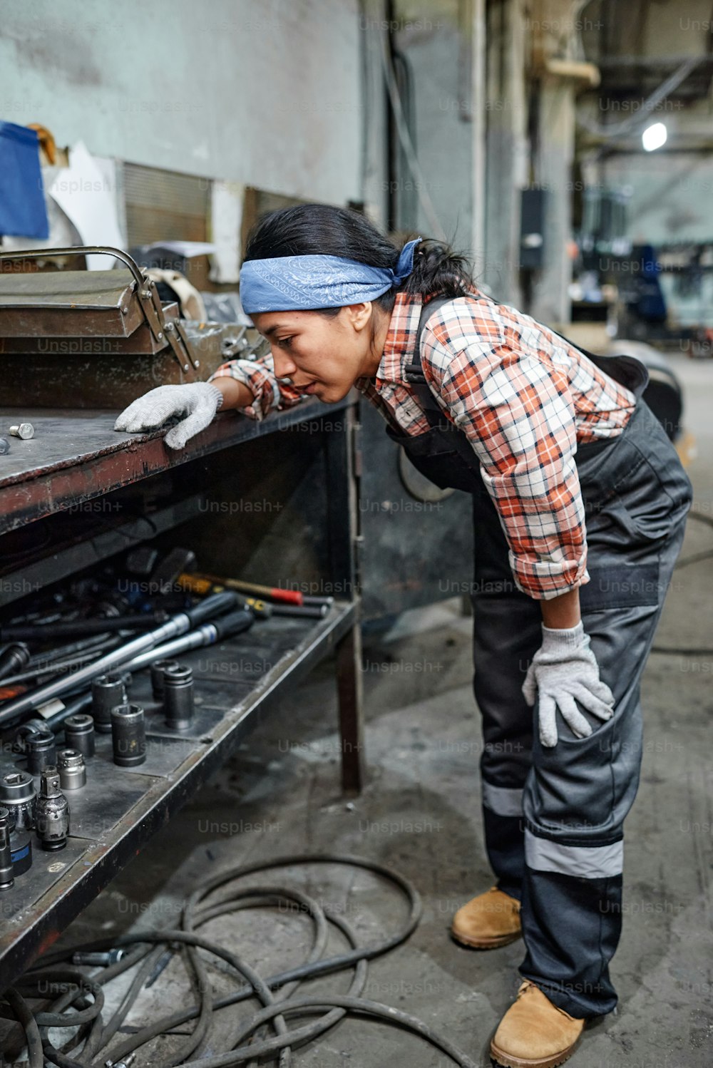 Young female technician in workwear bending in front of workbench while choosing handtools for repairing industrial machine