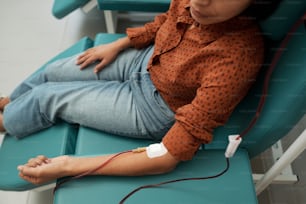 Young female giving her blood for sick patients with dropper tube connected to her vein while sitting in hospital ward