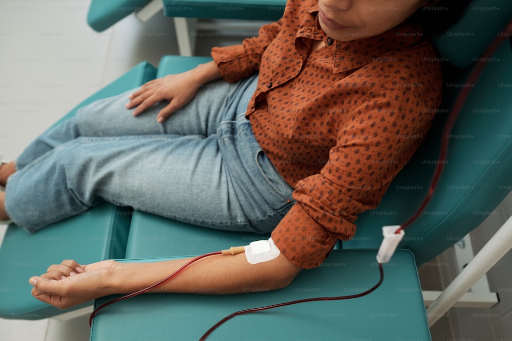 Young female giving her blood for sick patients with dropper tube connected to her vein while sitting in hospital ward