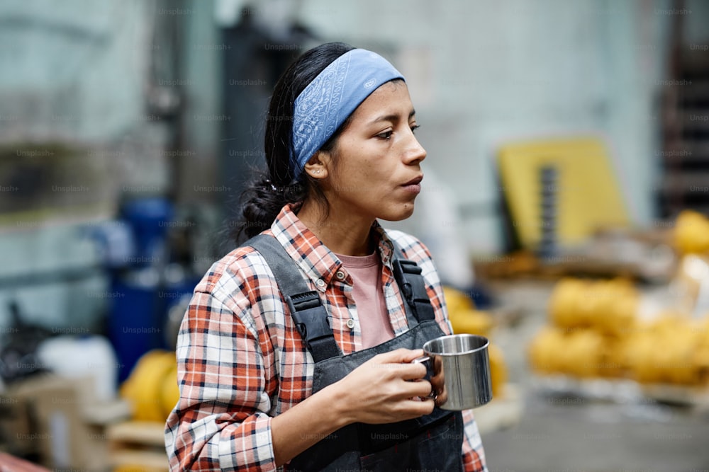 Young serious female builder in coveralls and blue headband holding mug with tea while having break in warehouse or workshop