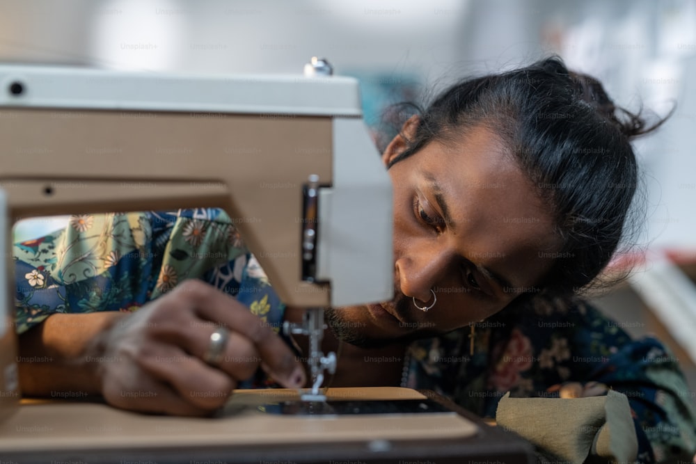 Serious guy of Indian ethnicity putting thread into needle eye of electric sewing machine while working over new fashion collection