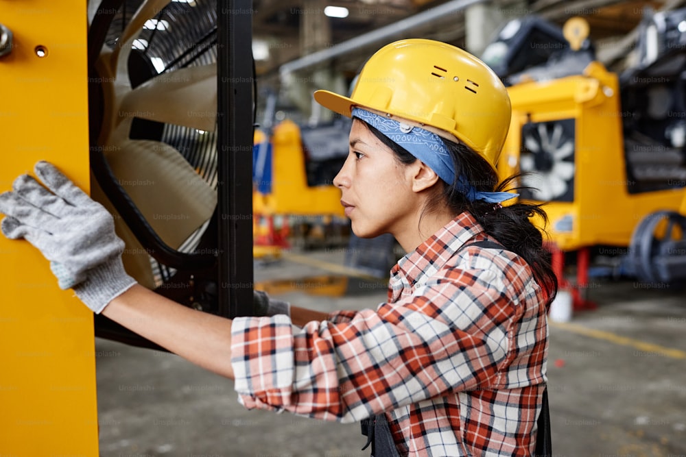 Young worker of modern factory in workwear and safety helmet checking motor of industrial machine while opening ventilated section