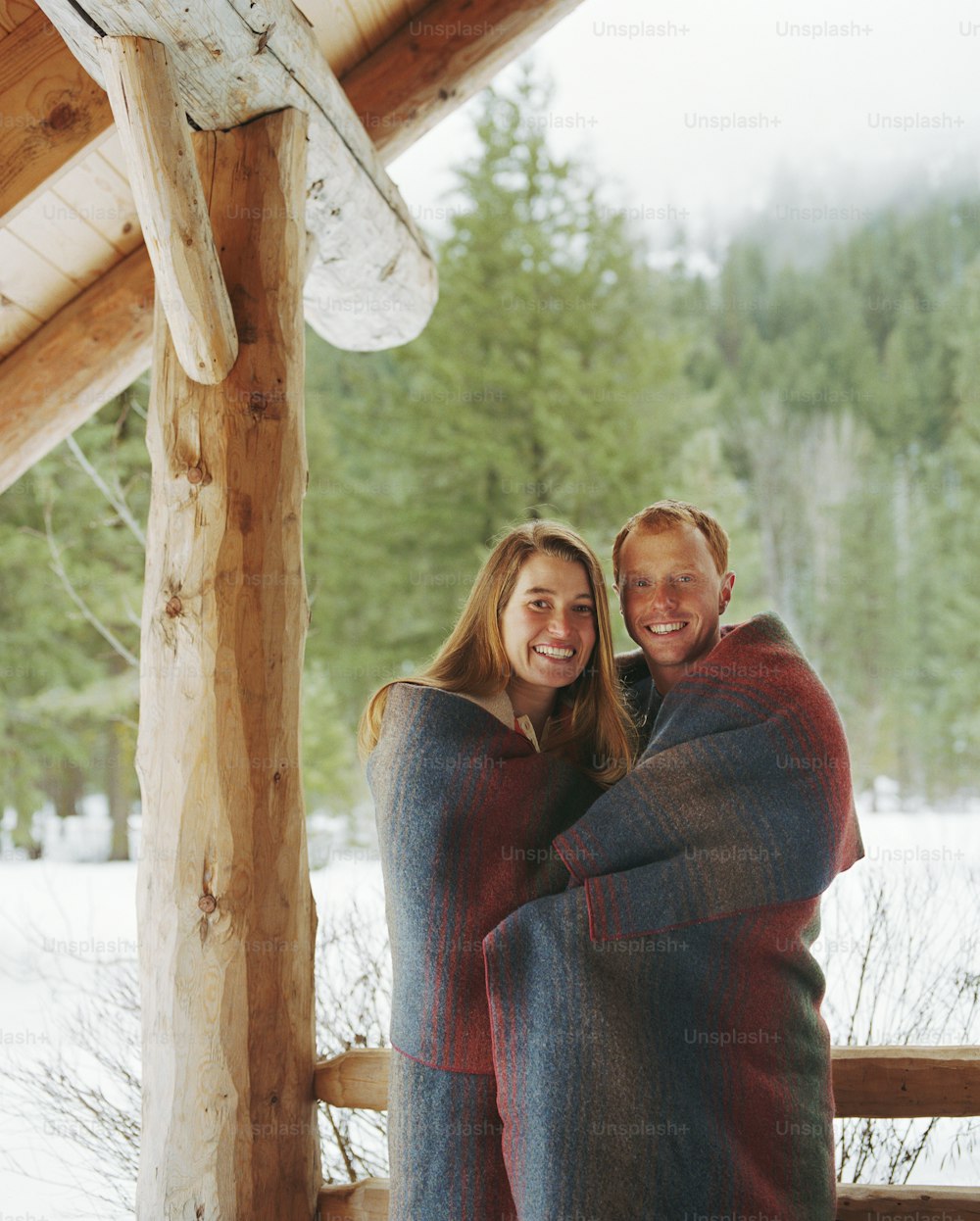Young couple wrapped in blanket on porch of cabin, smiling, portrait