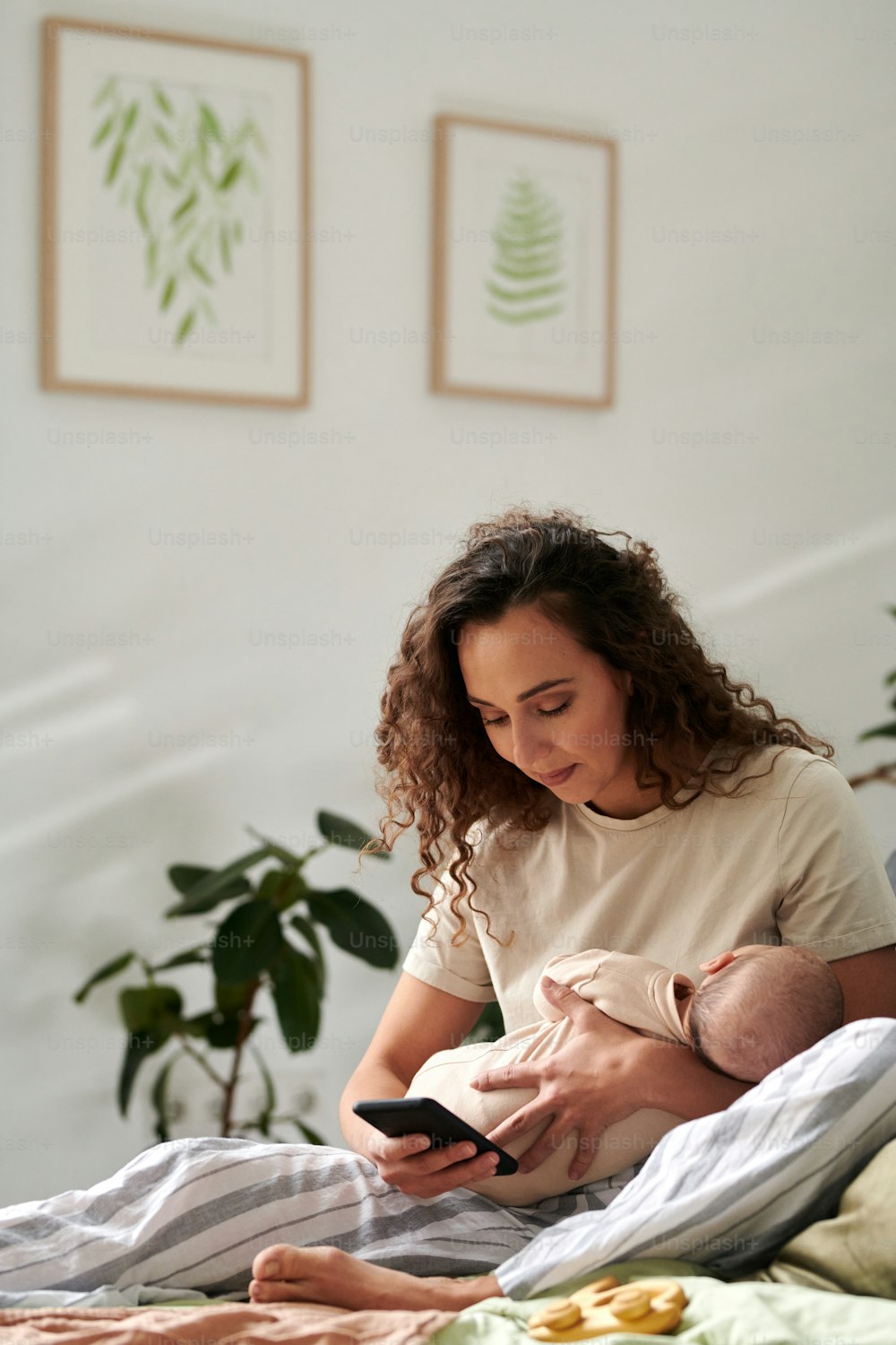 액자에 사진이 있는 벽에 편안한 더블 침대에 앉아 있는 동안 휴대 전화로 문자를 보내는 손에 아기를 둔 젊은 어머니