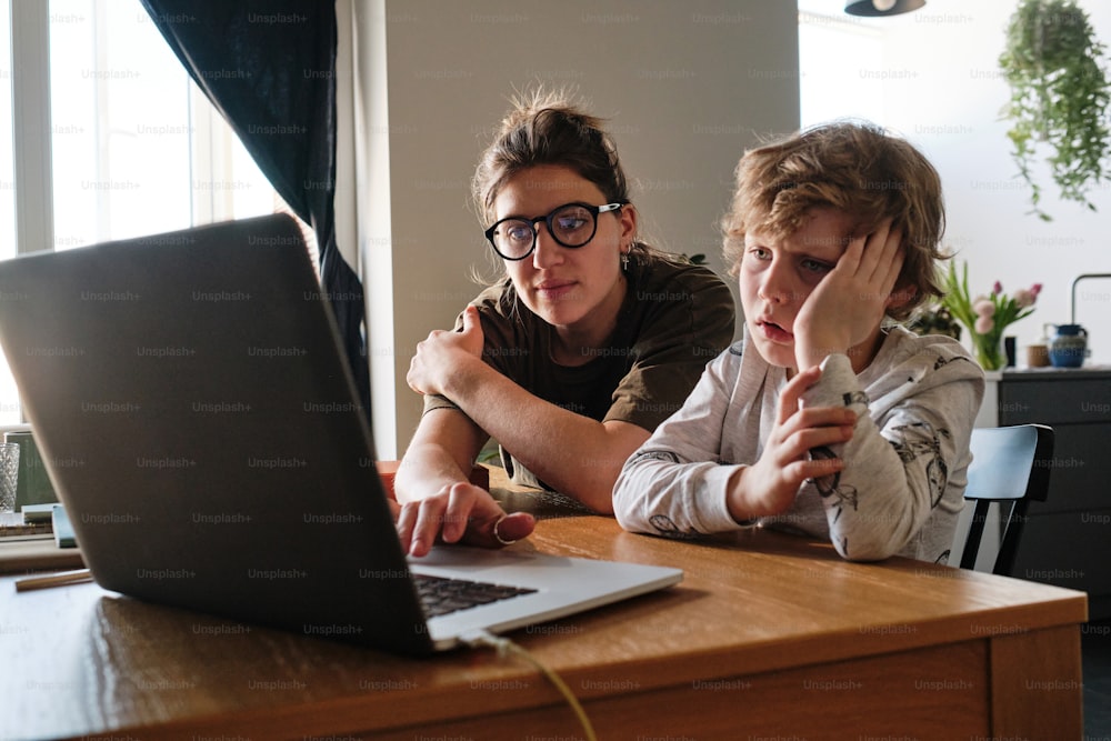 Little boy looking at monitor of laptop and thinking, he doing tasks online together with teacher at table