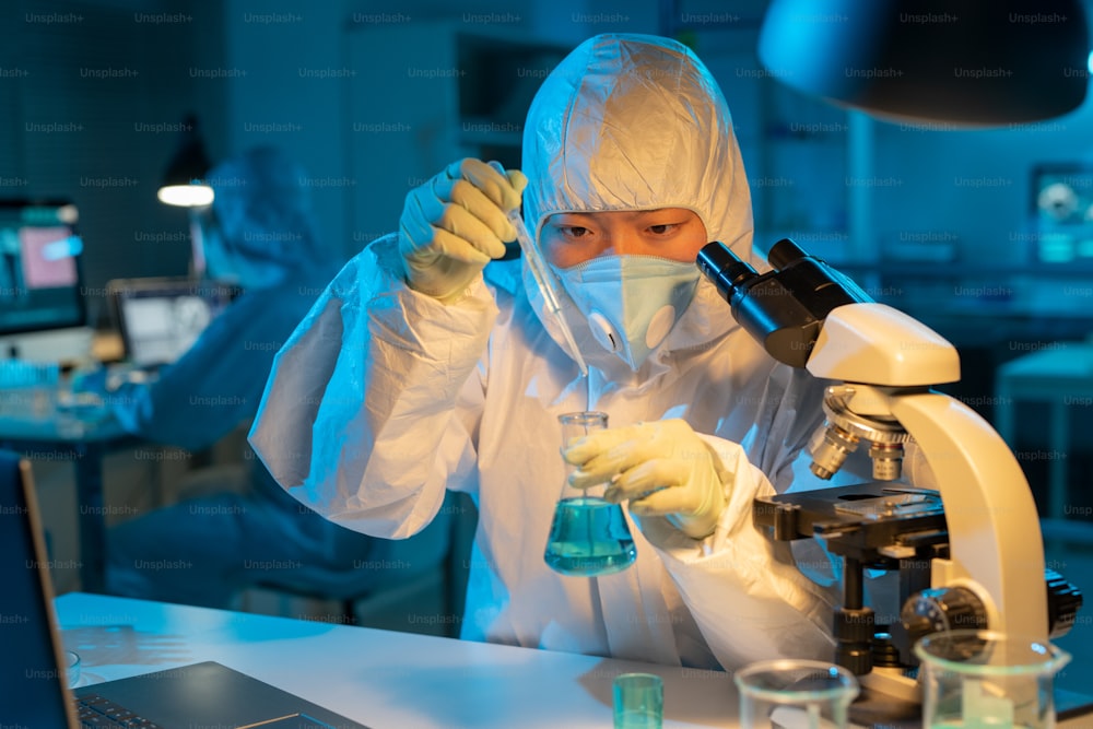 Investigadora o viróloga contemporánea en ropa de trabajo protectora que pone una gota de antídoto en un matraz con sustancia líquida