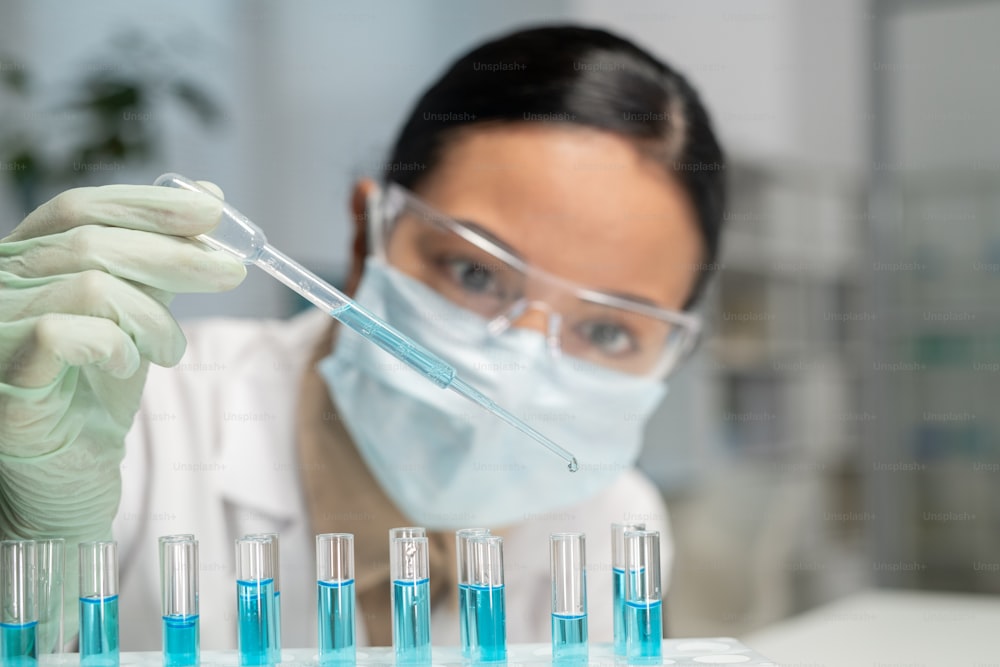 若い女性化学者またはウイルス学者が、青い液体が入ったいくつかのフラスコの1つに新しい化学物質を滴下します
