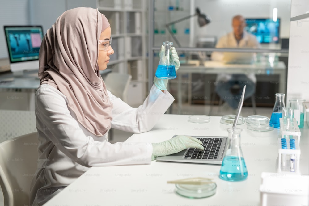 Seitenansicht einer jungen muslimischen Wissenschaftlerin im Hijab beim Betrachten des Reagenzglases mit blauer Flüssigkeit in der behandschuhten Hand während der wissenschaftlichen Forschung