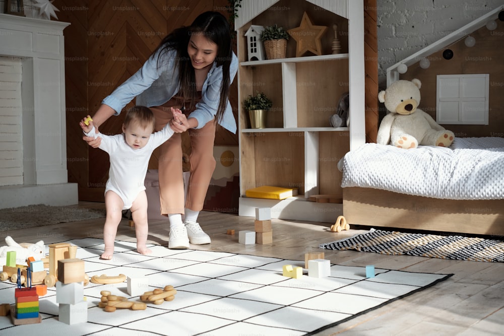 Cheerful young mother spending time at home teaching her lovely baby daughter how to walk